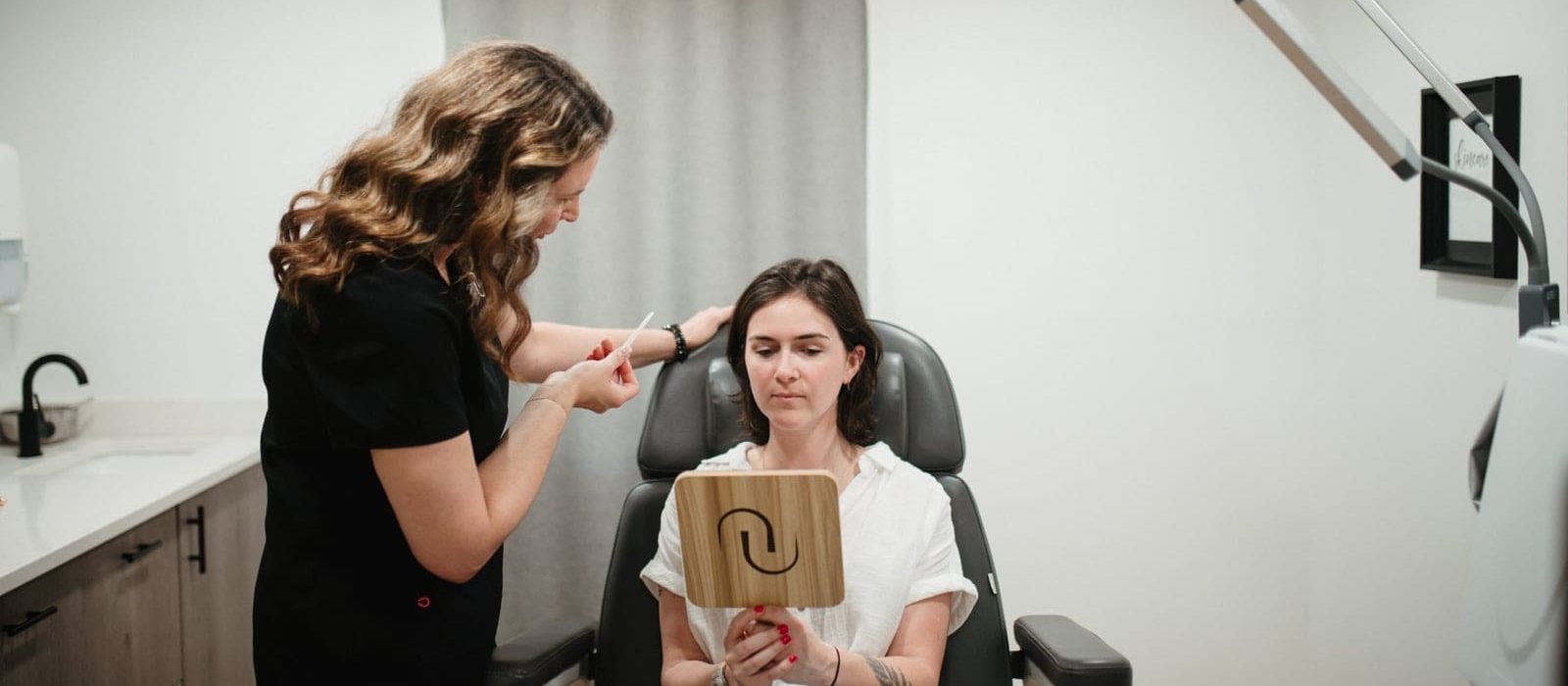 An esthetician consults a client holding a mirror in a medical spa treatment room.