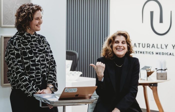Two women talking inside a medical spa with a logo on the wall.